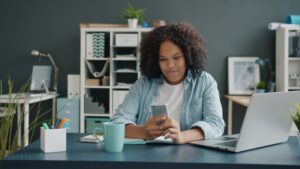 Young woman working in a modern office setting, actively using a smartphone at her desk.